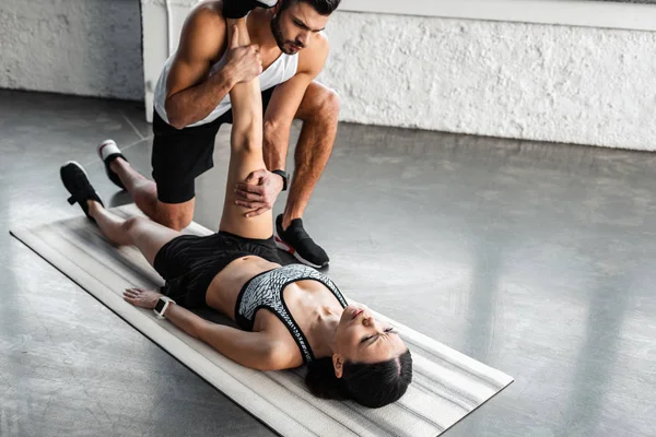 Athletic young man helping sporty woman stretching on yoga mat in gym — Stock Photo