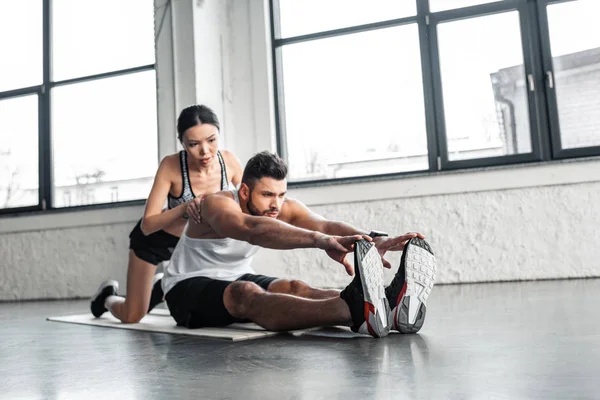 Atractiva joven deportiva ayudando a deportista muscular a estirarse en la esterilla de yoga en el gimnasio - foto de stock