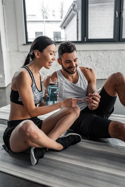 Lächelndes junges Paar in Sportbekleidung mit Smartphone, während es sich nach dem Training im Fitnessstudio auf Yogamatten ausruht — Stockfoto