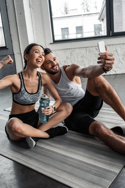 Glückliches sportliches junges Paar macht Selfie mit Smartphone, während es sich nach dem Training im Fitnessstudio auf Yogamatten ausruht — Stockfoto