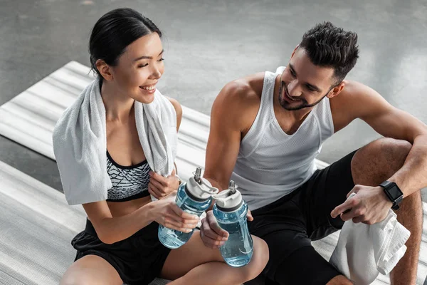 High angle view of smiling young couple in sportswear holding bottles of water while sitting and resting in gym — Stock Photo