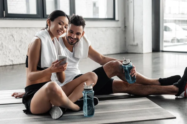 Щаслива спортивна молода пара відпочиває на йога килимках і використовує смартфон разом у спортзалі — стокове фото