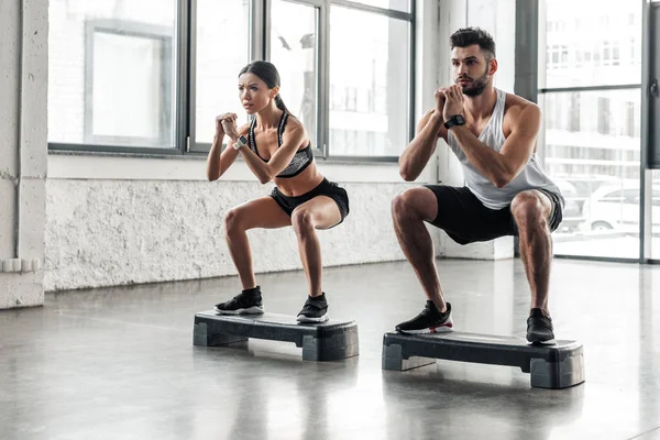 Concentrated athletic young couple in sportswear squatting on step platforms in gym — Stock Photo