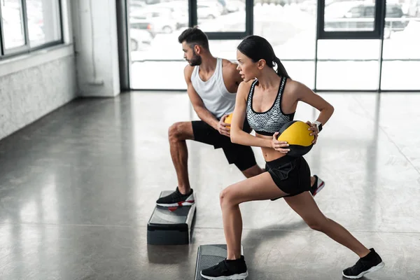 Vista lateral de una joven pareja deportiva sosteniendo pelotas de medicina y haciendo ejercicio en plataformas escalonadas en el gimnasio - foto de stock