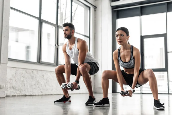 Sporty young man and woman squatting with dumbbells in gym — Stock Photo