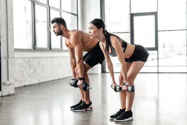 Vista lateral de la joven pareja deportiva entrenando con pesas y mirando hacia otro lado en el gimnasio - foto de stock
