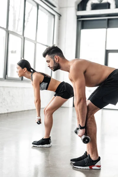 Vue latérale du jeune couple sportif faisant de l'exercice avec des haltères dans la salle de gym — Photo de stock