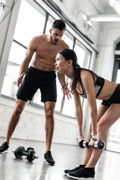 Allenatore di sesso maschile che parla con ragazza sportiva che si esercita con i manubri in palestra — Foto stock