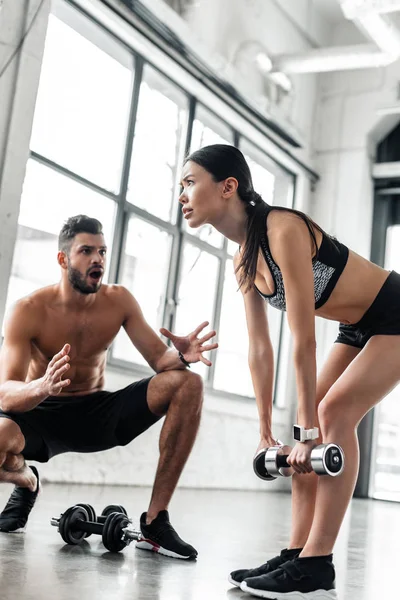 Masculino treinador gesticulando e falando com menina desportiva exercitando com halteres no ginásio — Fotografia de Stock