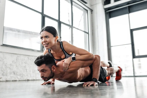 Musculoso sin camisa joven haciendo flexiones con chica deportiva en la espalda en el gimnasio - foto de stock