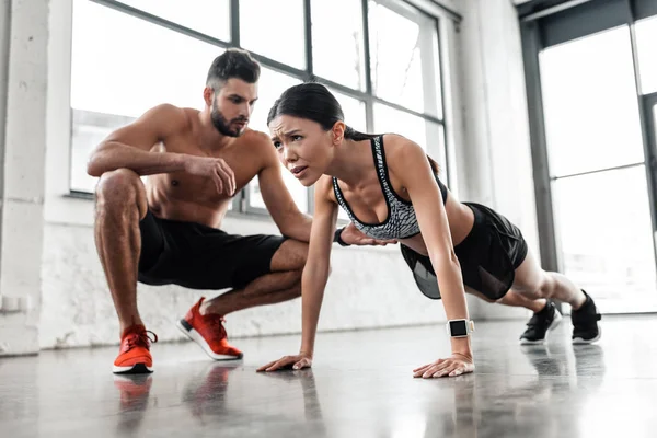 Muskulöse hemdlose männliche Trainerin schaut junge Sportlerin beim Liegestütz im Fitnessstudio an — Stockfoto