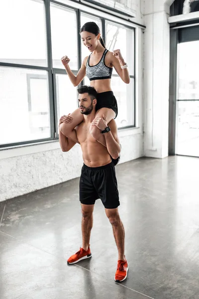 Schöner muskulöser Mann ohne Hemd trägt fröhliches sportliches Mädchen auf Schultern in der Turnhalle — Stockfoto