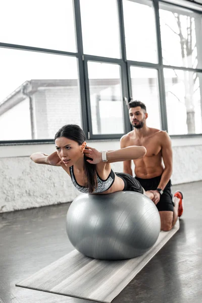 Muscular shirtless trainer helping young sportswoman exercising on fit ball in gym — Stock Photo