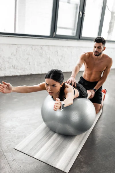 Hemdlose männliche Trainerin hilft sportlichem Mädchen beim Training auf Fitnessball im Fitnessstudio — Stockfoto