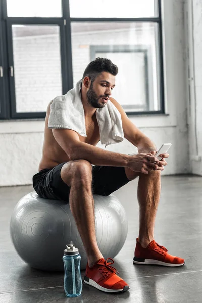 Молодий м'язистий босоніж спортсмен сидить на м'ячі і використовує смартфон у тренажерному залі — стокове фото