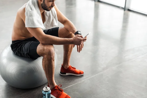 Tiro cortado de homem nu-peito muscular sentado na bola em forma e usando smartphone no ginásio — Fotografia de Stock