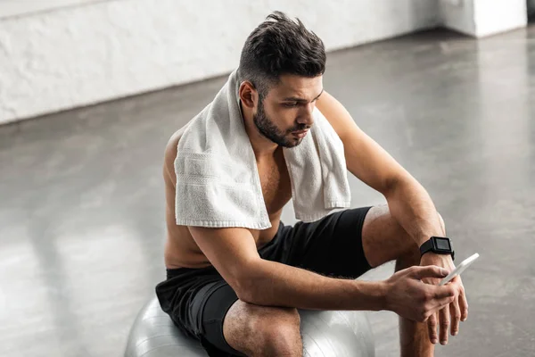 Hochwinkelaufnahme eines muskulösen Mannes mit nacktem Oberkörper, der auf einem fitten Ball sitzt und das Smartphone im Fitnessstudio benutzt — Stockfoto