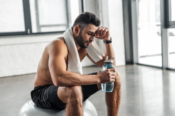Уставший мускулистый спортсмен с полотенцем и бутылкой воды, сидящий на фитнес-мяче в тренажерном зале — стоковое фото