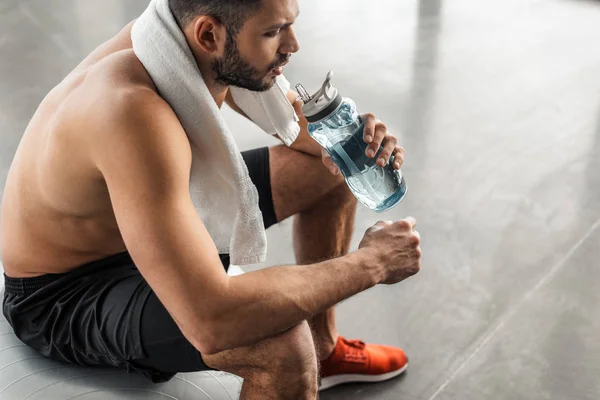 Schnappschuss eines jungen Sportlers mit nacktem Oberkörper, der mit Handtuch auf einem Ball sitzt und in der Turnhalle Wasser trinkt — Stockfoto