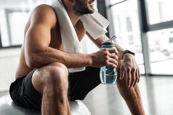 Tiro recortado de deportista sin camisa con toalla y botella de agua sentado en la bola en forma en el gimnasio - foto de stock