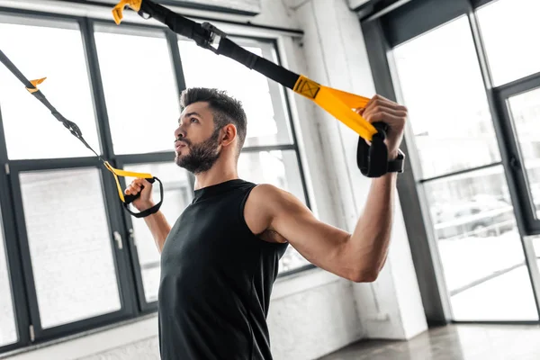 Athletic young man in sportswear training with suspension straps and looking up in gym — Stock Photo