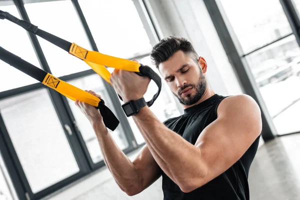 Schöner muskulöser junger Mann, der beim Training mit Aufhängebändern im Fitnessstudio auf den Bizeps schaut — Stockfoto