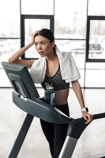 Tired young sportswoman with towel standing on treadmill and looking away in gym — Stock Photo