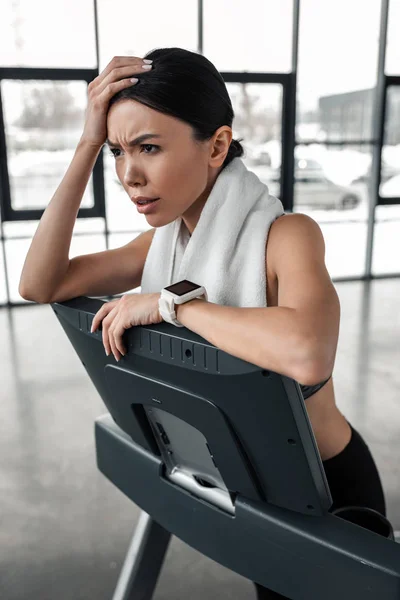 Exhausted young sportswoman leaning at treadmill after workout and looking away in gym — Stock Photo