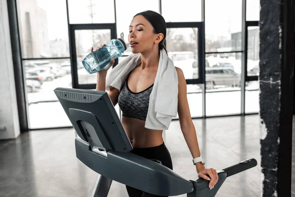 Beautiful young sportswoman with towel on shoulders standing on treadmill and drinking water in gym — Stock Photo
