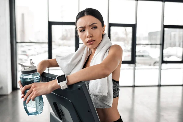 Müde junge Sportlerin mit Handtuch und Wasserflasche auf Laufband stehend und im Fitnessstudio wegschauend — Stockfoto