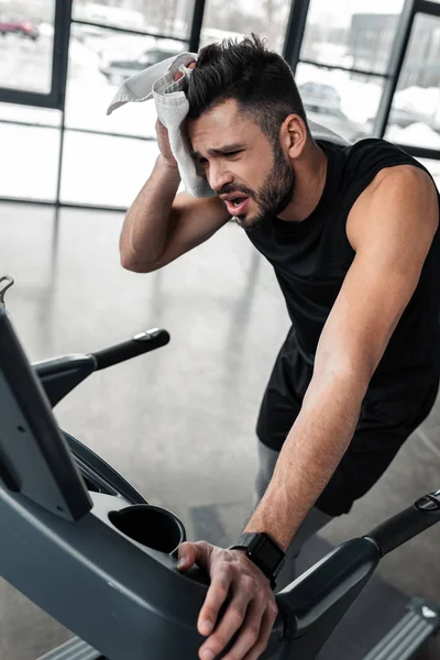 Tired young sportsman training on treadmill and wiping sweat with towel in gym — Stock Photo