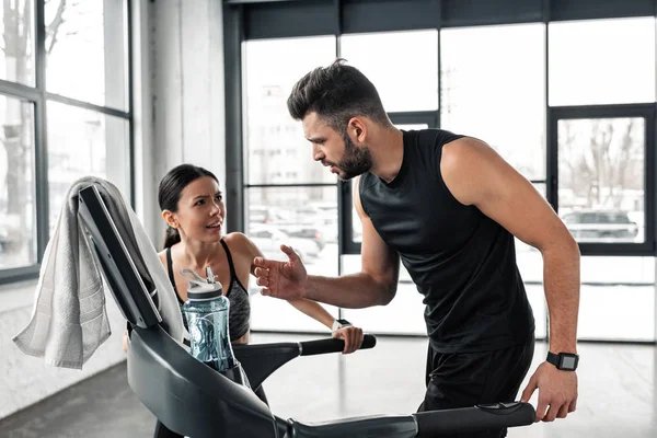 Young sportswoman looking at exhausted athletic man running on treadmill in gym — Stock Photo