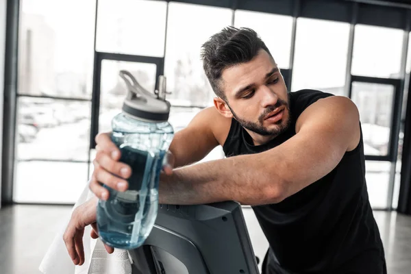 Joven deportista cansado apoyado en la cinta de correr y sosteniendo la botella de deportes con agua en el gimnasio - foto de stock