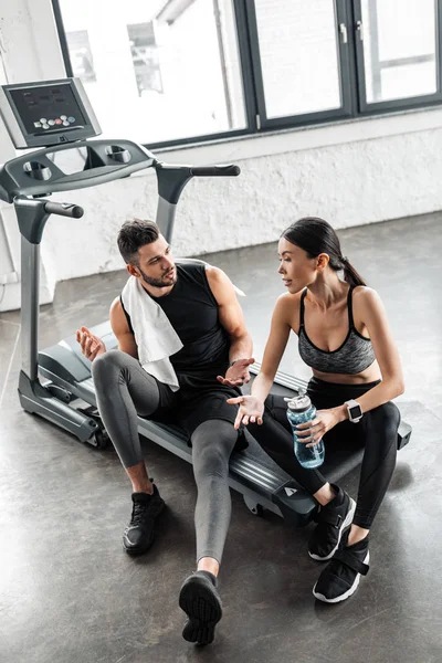 Vue grand angle de jeune couple sportif avec serviette et bouteille d'eau parlant tout en étant assis sur le tapis roulant après l'entraînement dans la salle de gym — Photo de stock