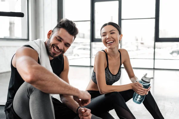 Alegre joven pareja riendo mientras se sienta en la cinta de correr y descansando después del entrenamiento en el gimnasio - foto de stock