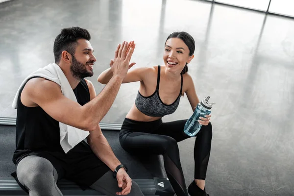 Hochwinkelaufnahme eines glücklichen sportlichen jungen Paares, das High Five gibt, während es nach dem Training im Fitnessstudio auf dem Laufband sitzt — Stockfoto