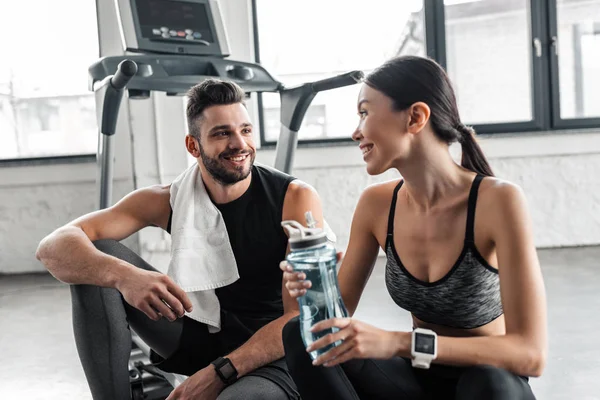 Smiling young couple with towel and bottle of water resting together on treadmill in gym — Stock Photo