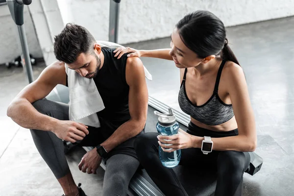 Vista de ángulo alto de pareja joven y cansada con toalla y botella de agua descansando juntos en la cinta de correr en el gimnasio - foto de stock