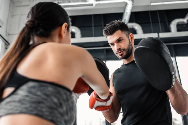Low angle view of male trainer looking at young sportswoman boxing in gym — Stock Photo