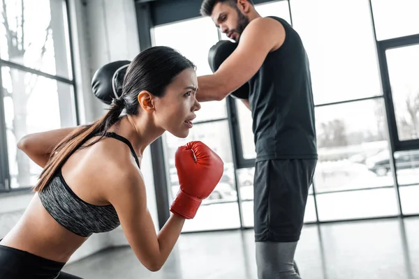 Joven deportista concentrada en guantes de boxeo haciendo ejercicio con entrenador en gimnasio - foto de stock