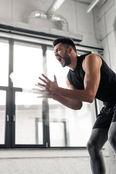 Vue à faible angle du sportif agressif criant et appliquant de la poudre de talc sur les mains dans la salle de gym — Photo de stock