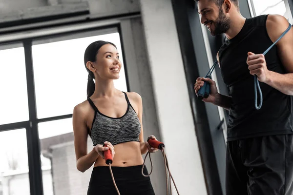 Happy sporty young couple holding jumping ropes and smiling each other in gym — Stock Photo