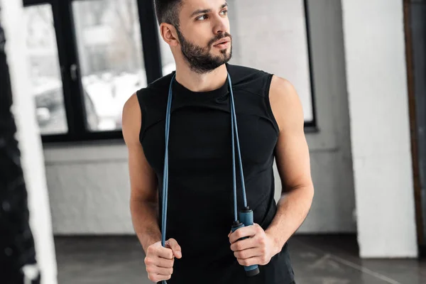 Cropped shot of handsome sportsman holding skipping rope around neck and looking away in gym — Stock Photo