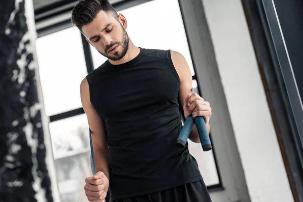 Низкий угол зрения на мускулистого молодого человека в спортивной одежде, держащего скакалку в спортзале — стоковое фото