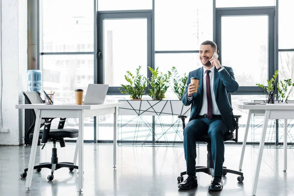 Alegre hombre de negocios sosteniendo taza de papel mientras habla en el teléfono inteligente en la oficina moderna - foto de stock