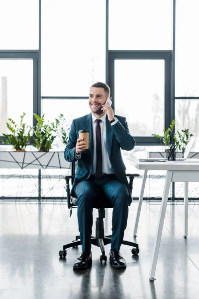 Homme d'affaires joyeux tenant tasse en papier et parler sur smartphone dans le bureau moderne — Photo de stock