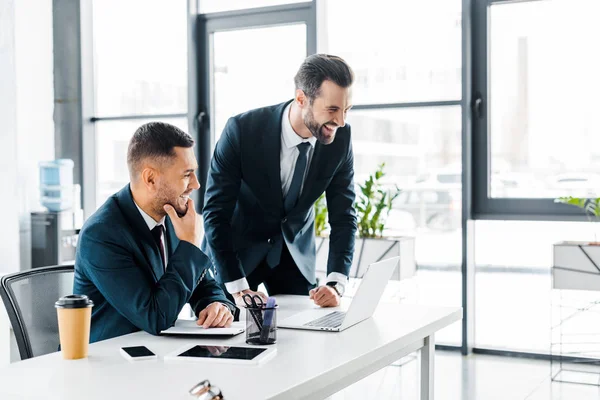 Hombres de negocios guapos riendo cerca de la computadora portátil en la oficina moderna — Stock Photo