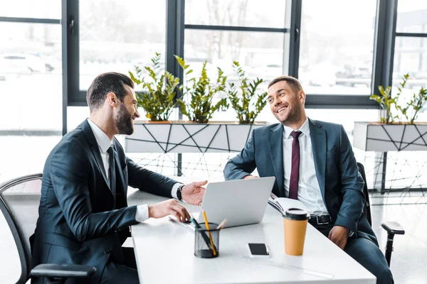 Guapo hombre de negocios mirando a su compañero de trabajo y sonriendo en la oficina moderna - foto de stock