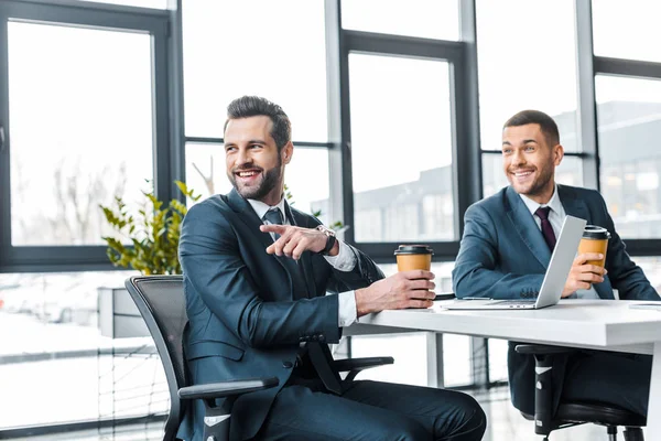 Веселый бизнесмен показывает пальцем, держа бумажную чашку рядом с коллегой в современном офисе — стоковое фото