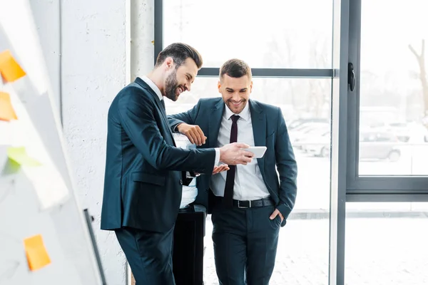 Hombre de negocios guapo mirando el teléfono inteligente con colega de pie con la mano en el bolsillo - foto de stock